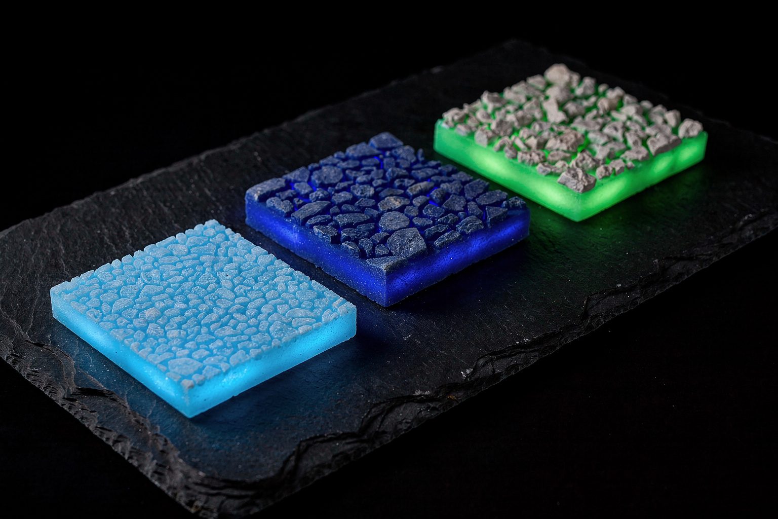 Illustration: three flat glowing path coating sample tiles in sky blue, deep blue and green, with fine to coarse stone chips